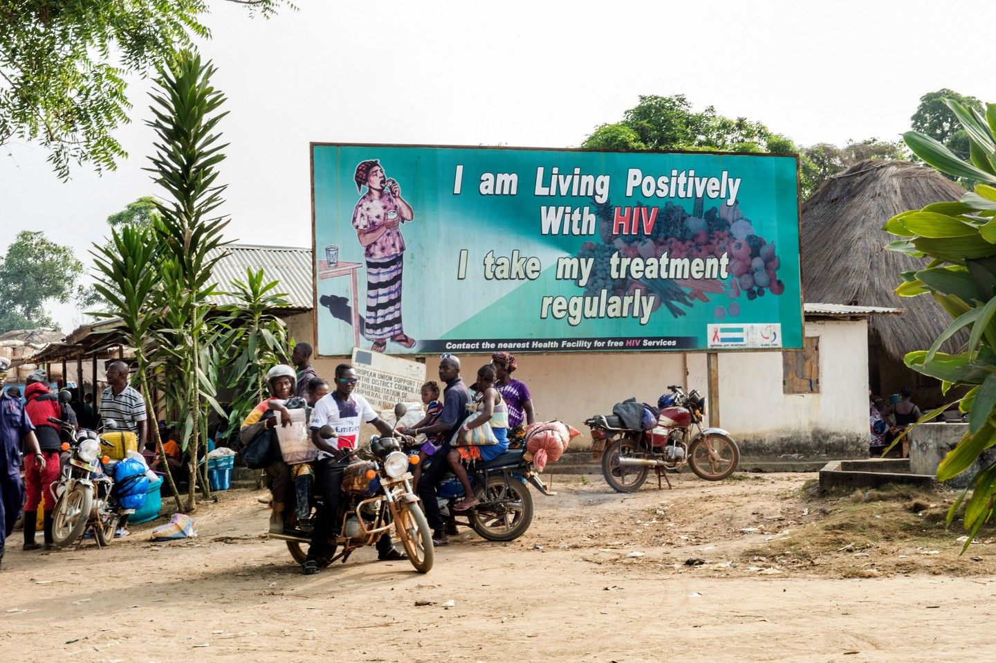 community members congregating outside next to a billboard with a positive message about living with HIV