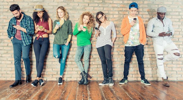 Nine young adults leaning against a wall checking their smart phones.